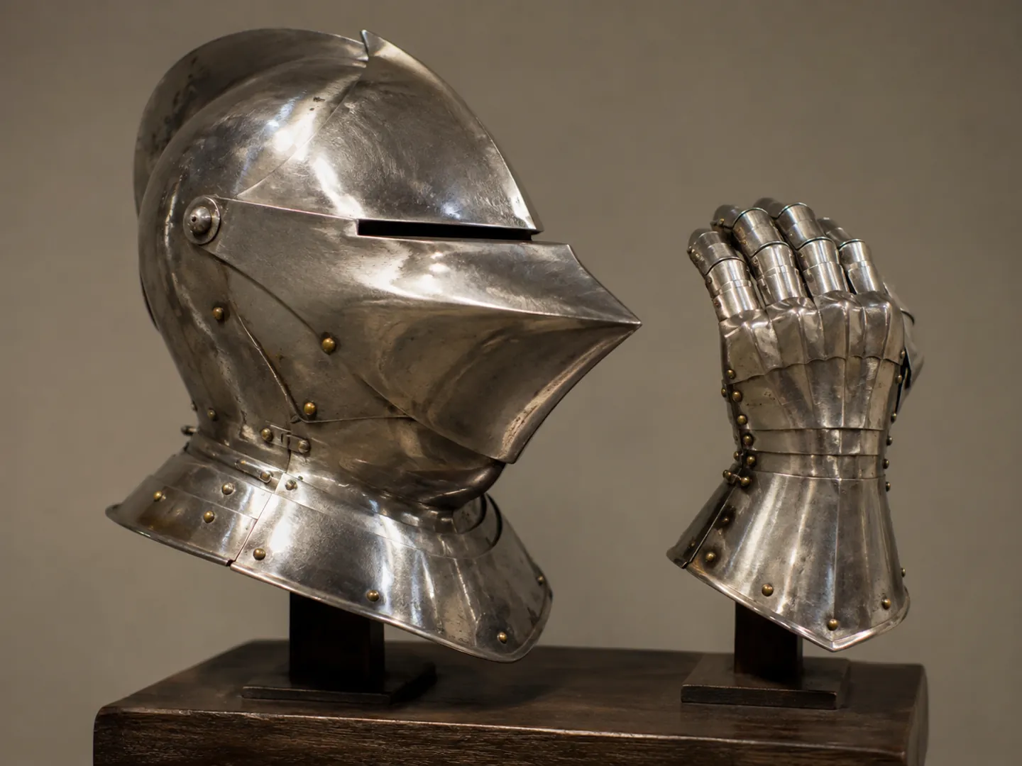 A steel jousting helmet with a narrow eye slit and a heavy gauntlet displayed on a wooden stand under soft museum light